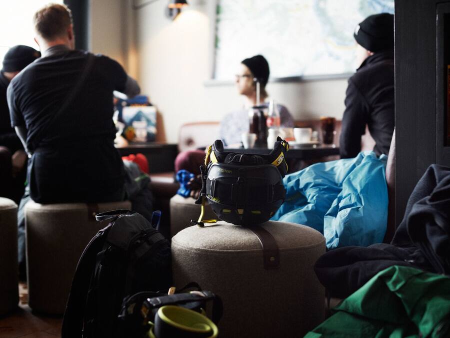 Eine Gruppe von Personen sitzt an einem Tisch in einem Raum mit Gepäck, was auf einen Wartebereich oder eine Lounge hindeutet.