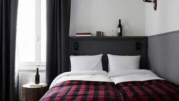 Una cama bien hecha con una colcha de cuadros rojos y negros, una mesita de madera pequeña con una botella de vino y una copa, y una estantería con libros y otra copa de vino encima de la cama.