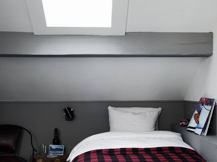 Ein Hotelzimmer mit Bett, einem kleinen Holfnachtkästchen, einem Sessel, einem Nachttisch, einem an der Wand befestigten Telefon und einem Fenster mit Blick zum Himmel.
