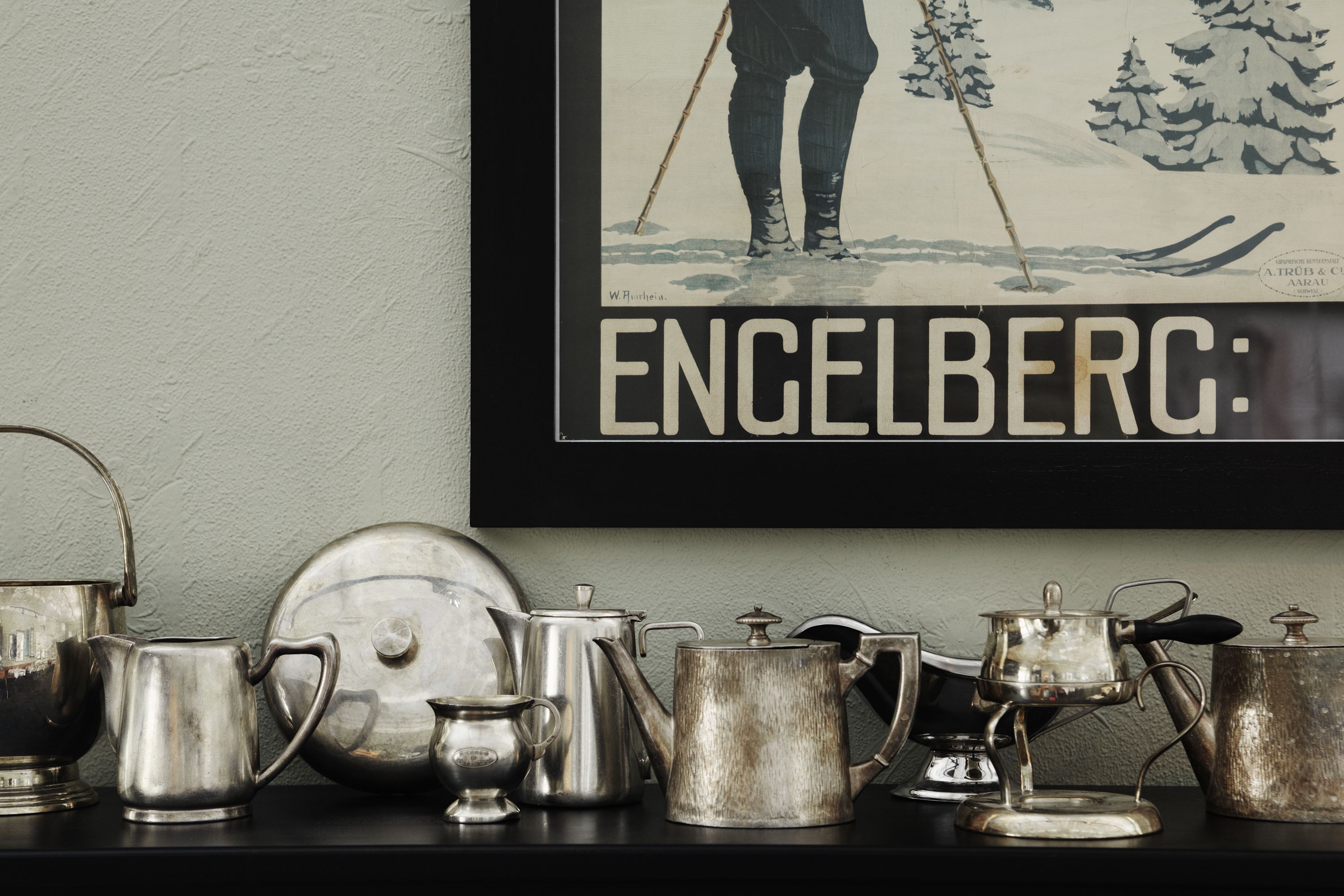 A collection of silver kitchenware on a shelf, including a kettle, teapot, and a large pot, with a framed picture of a skier above them.