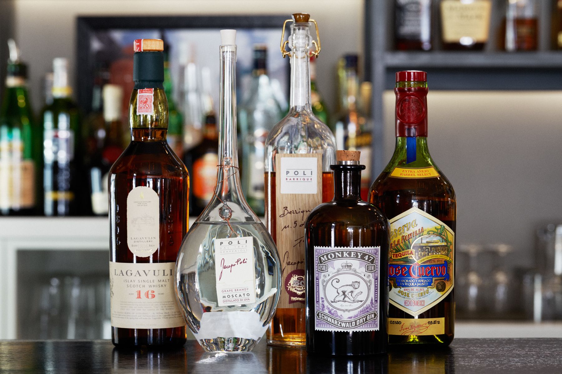Verschillende flessen op een barkruk, waaronder Lagavulin Schotse Whisky, een heldere fles met een kurk, en een fles Monkey 47.