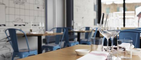 A dining table with a white napkin, silverware, and clear glasses, set against a backdrop of blue chairs and a wall with architectural drawings.