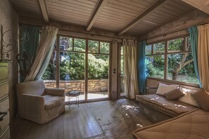 Una habitación acogedora con techo de madera, un sofá, una silla y una mesita. Amplias ventanas ofrecen vistas a zonas verdes.