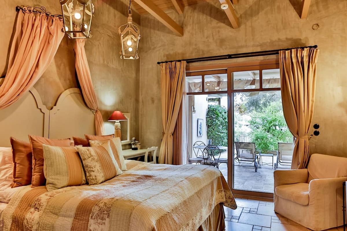 A bedroom with a large bed, a desk, and a chair, with a view of an outdoor patio.