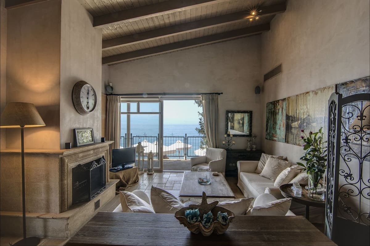 A spacious living room with a fireplace, comfortable seating, and a large window offering a view of the sea.