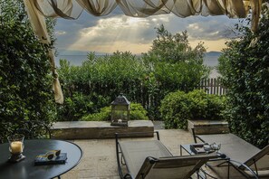 Un patio al aire libre con vistas al mar, que incluye una mesa con una vela, vasos y un libro, rodeado de sillas y un farol.