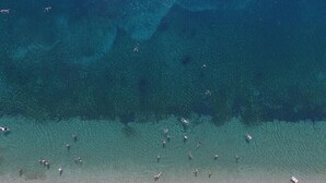 An aerial view of a beach with clear shallow waters, a row of sun loungers, and people swimming and sunbathing. - Six Keys Seaside Lodge (South Pelion)