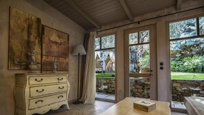 A room with a rustic wooden dresser, a table with a box, and a view of a garden through large windows. - Six Keys Seaside Lodge (South Pelion)
