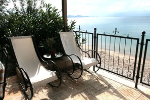 Two white cushioned chairs with metal frames, a small round table, and a potted plant on a balcony with a view of a beach and the sea. - Six Keys Seaside Lodge (South Pelion)