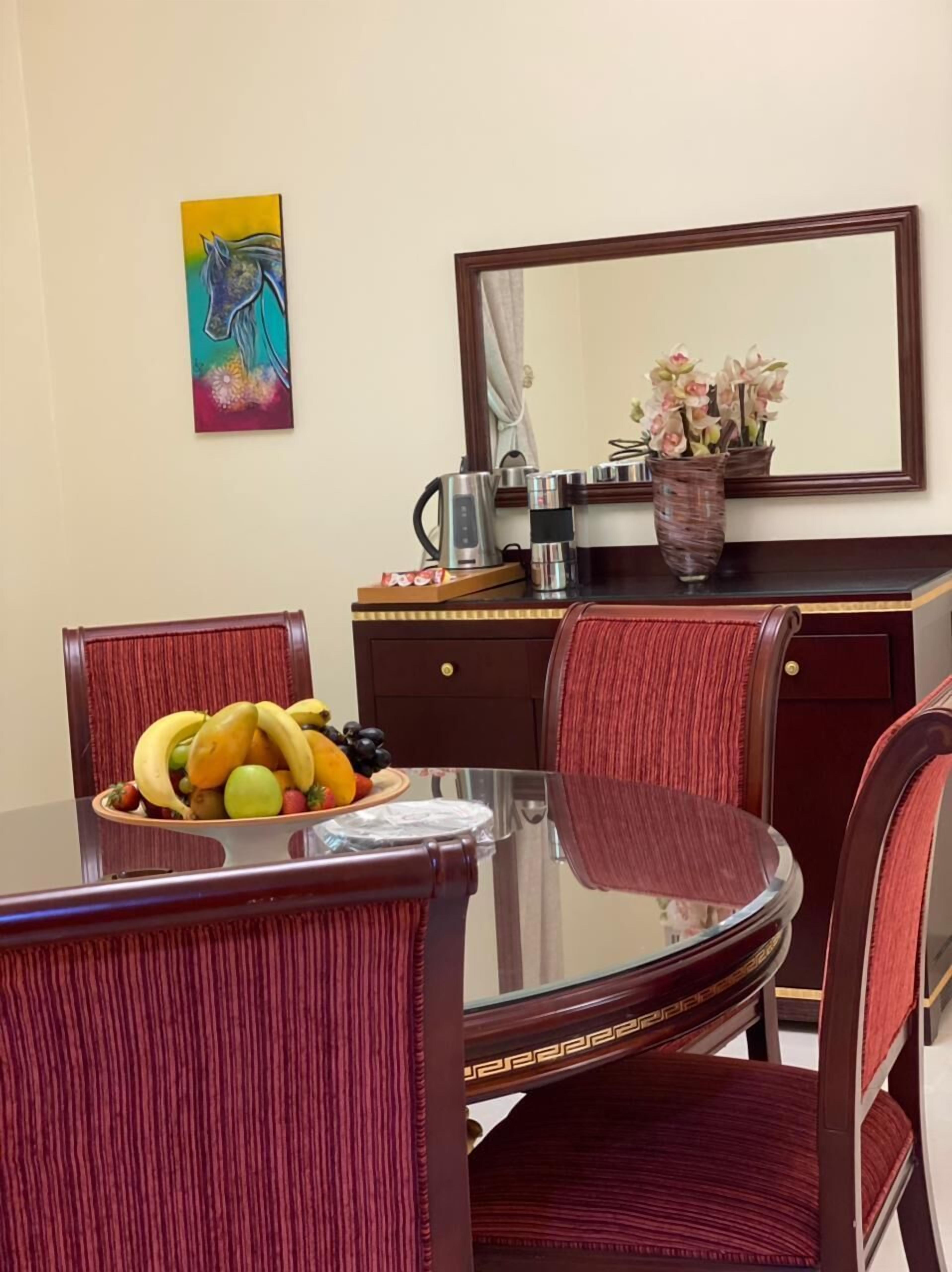 Un comedor con una mesa de vidrio, sillas tapizadas en rojo, un frutero, una cafetera, un jarrón con flores y un cuadro en la pared.
