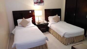 Habitación de hotel con dos camas, cada una con ropa blanca y falda color beige. Entre las camas hay una mesita de noche con una lámpara y un teléfono. Un cuadro cuelga de la pared sobre las camas.