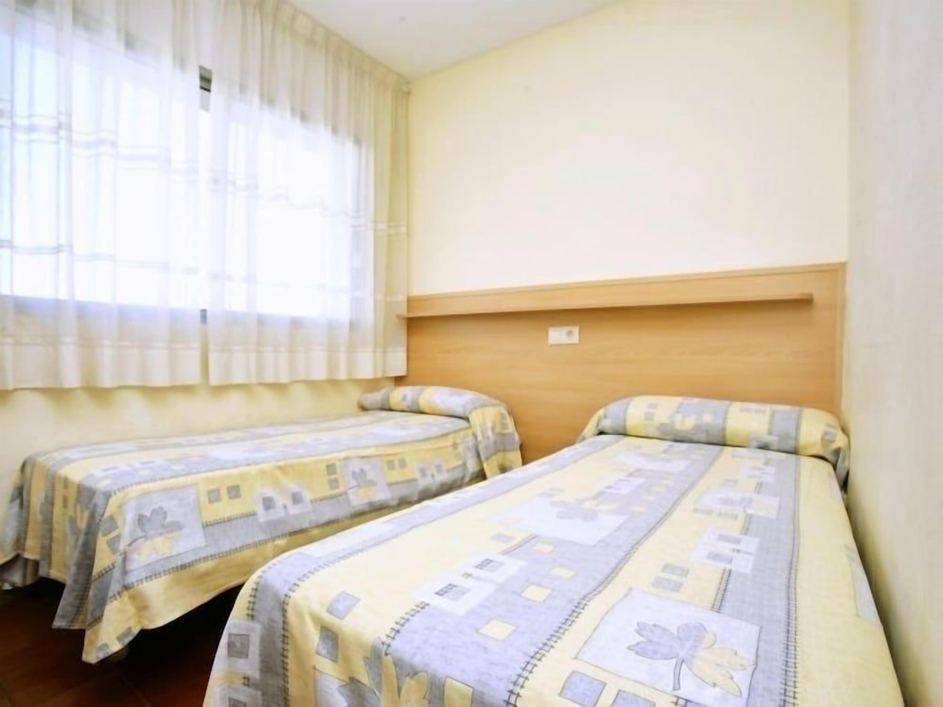Habitación con dos camas, cada una con una colcha estampada, cabecero de madera y ventana con cortinas de encaje.