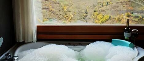 A bathtub filled with bubbles and a bottle of wine, with a view of a mountainous landscape through a window.