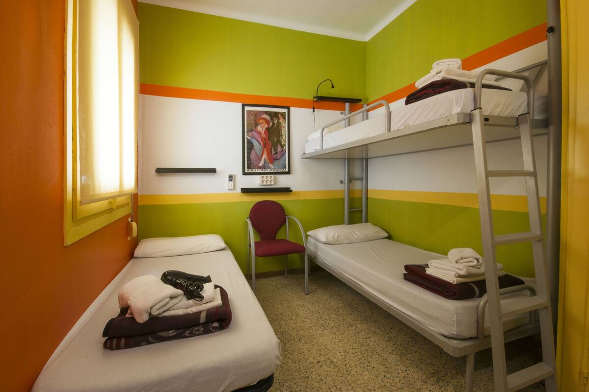 A dormitory room with bunk beds, a chair, a small table, and a poster on the wall.