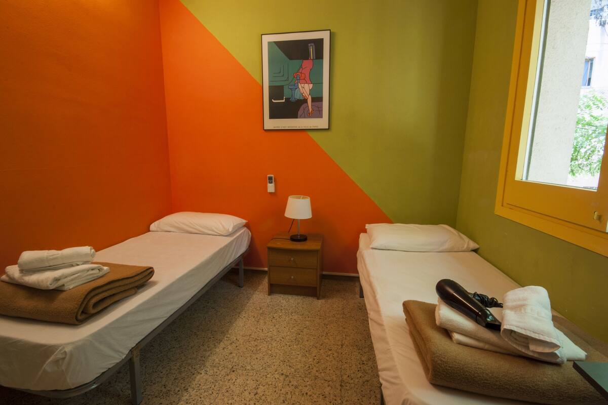 A small hotel room with two single beds, a nightstand with a lamp, and a window with a view.