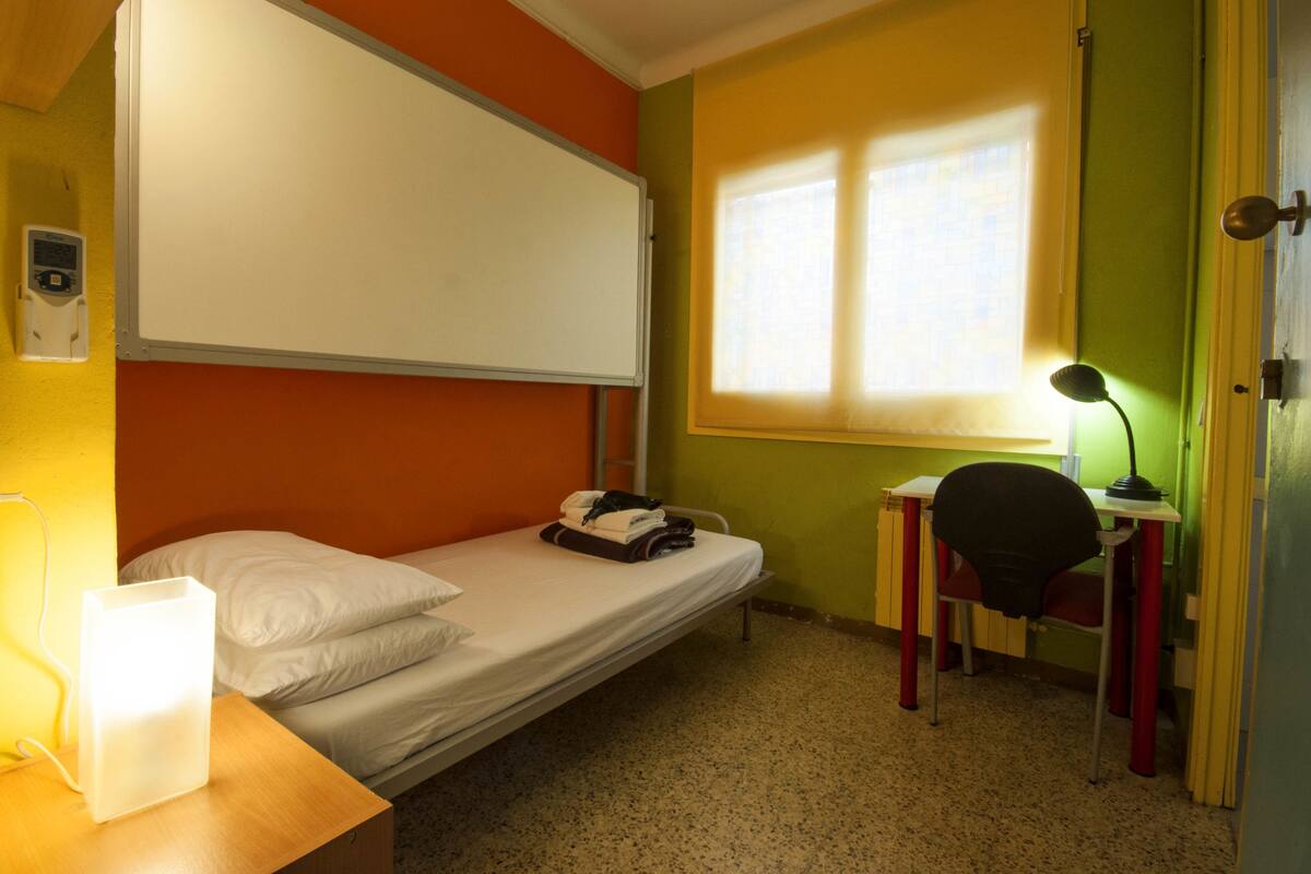 A small, single-bed room with a white bed, a desk with a chair, a window with blinds, and a wall-mounted light.