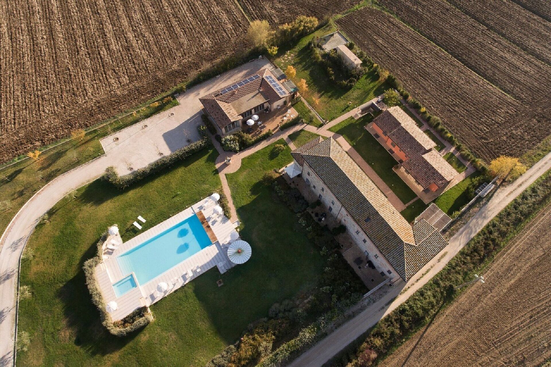 An aerial view of a property with a swimming pool, a large building with a tiled roof, and surrounding fields.