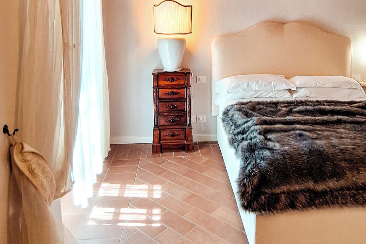 A bedroom with a wooden ceiling, a bed with a white headboard, a nightstand with a lamp, and a window with curtains.