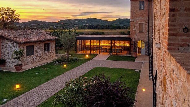 A courtyard with a brick pathway, a stone building, and a glass structure with warm lighting. - Anna Boccali Resort (Corciano)