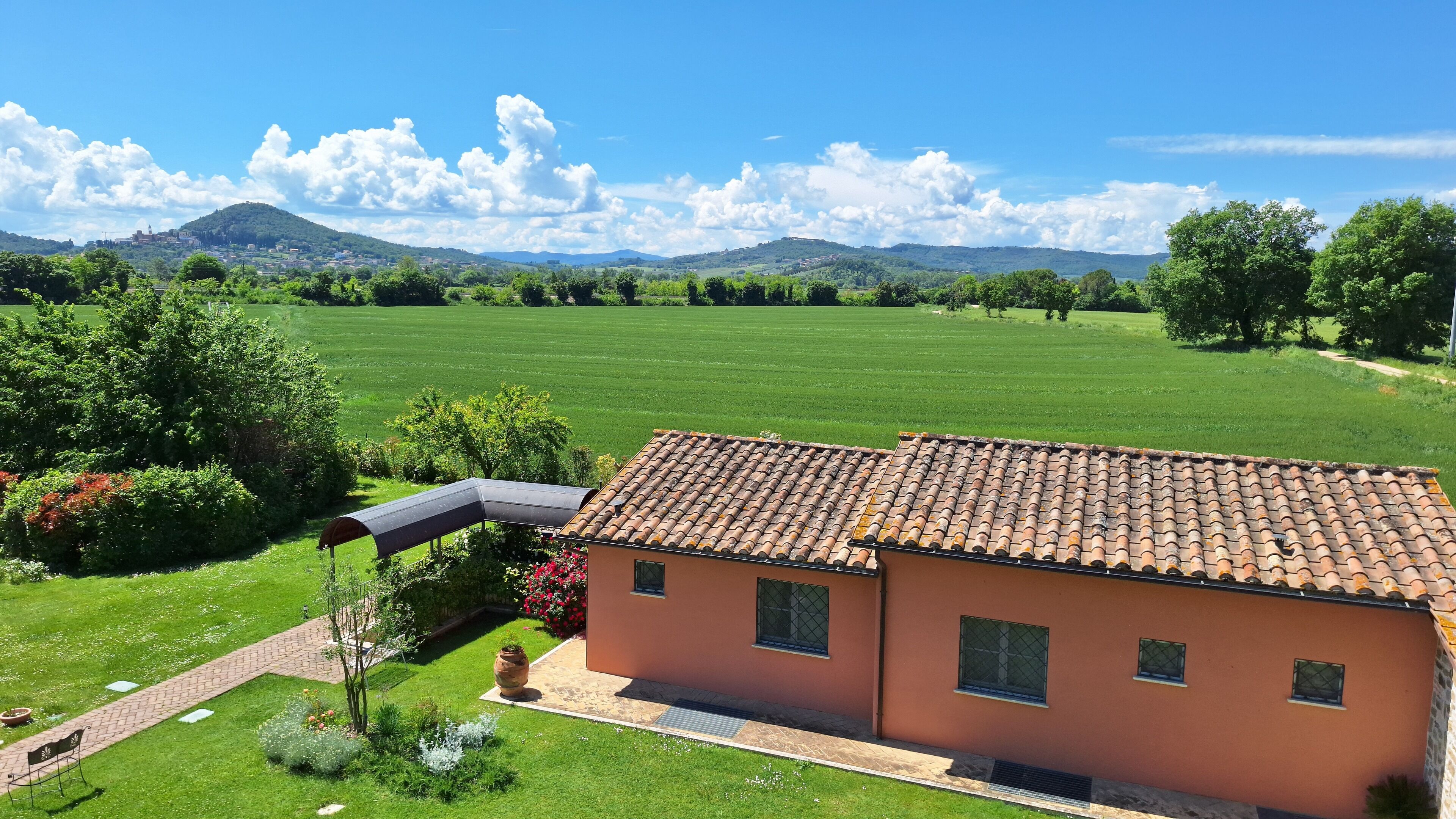 Et hus med tegltag, omgivet af en velholdt græsplæne og have, med udsigt til fjerne bakker og en klar himmel.