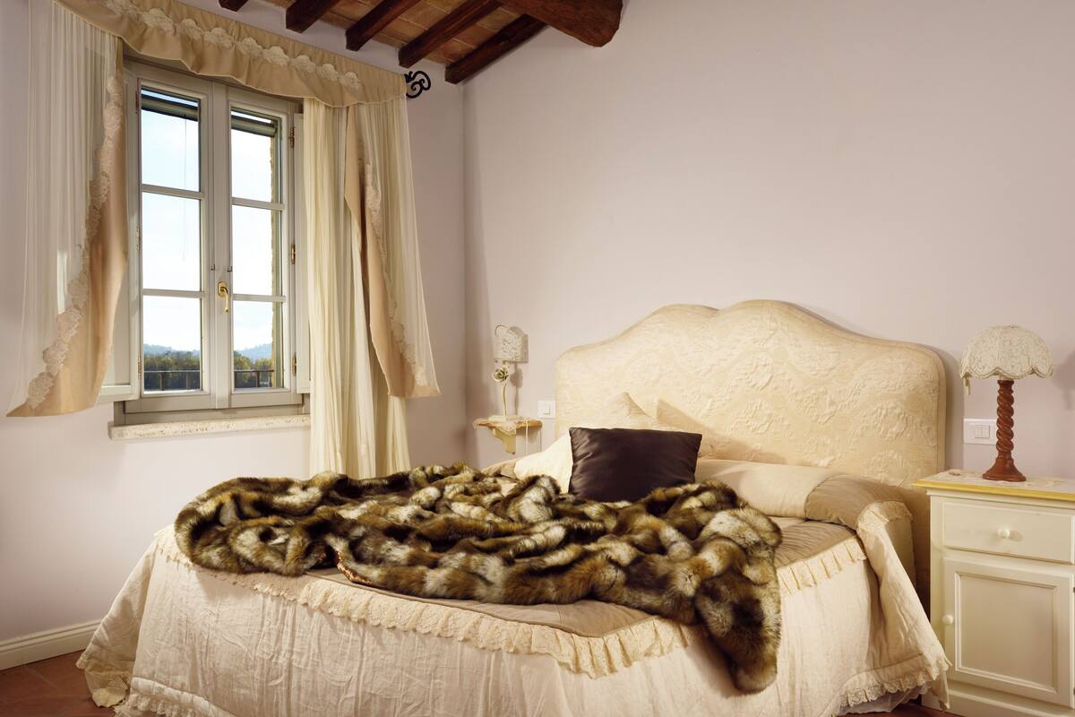 A bedroom with a bed covered in a fur blanket, a window with curtains, a bedside table with a lamp, and a nightstand with a mirror.