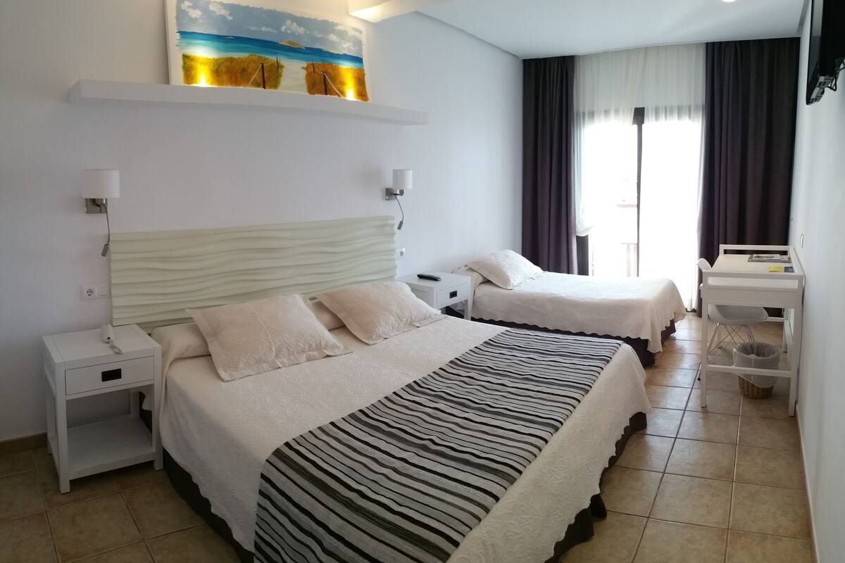 Ein Hotelzimmer mit zwei Betten, einem Schreibtisch, einem Sessel, einem Nachttisch, einer Lampe, einem Gemälde und einem fenster mit Vorhängen.