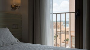 Une chambre d’hôtel avec un lit, une table de chevet, une lampe fixée au mur et une vue sur la ville par une fenêtre munie de rideaux.