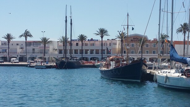 Un port de plaisance avec plusieurs bateaux amarrés, un bâtiment portant l’inscription « HÔTEL & BAHIA », et des palmiers en arrière-plan.