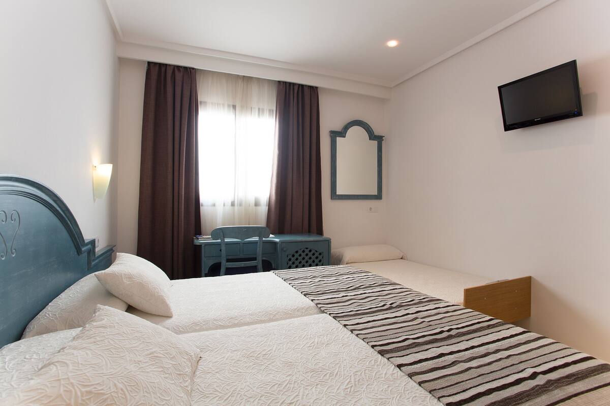 Ein Hotelzimmer mit zwei Betten, einem Schreibtisch, einem Stuhl, einem Fernseher und einem Fenster mit Vorhängen.