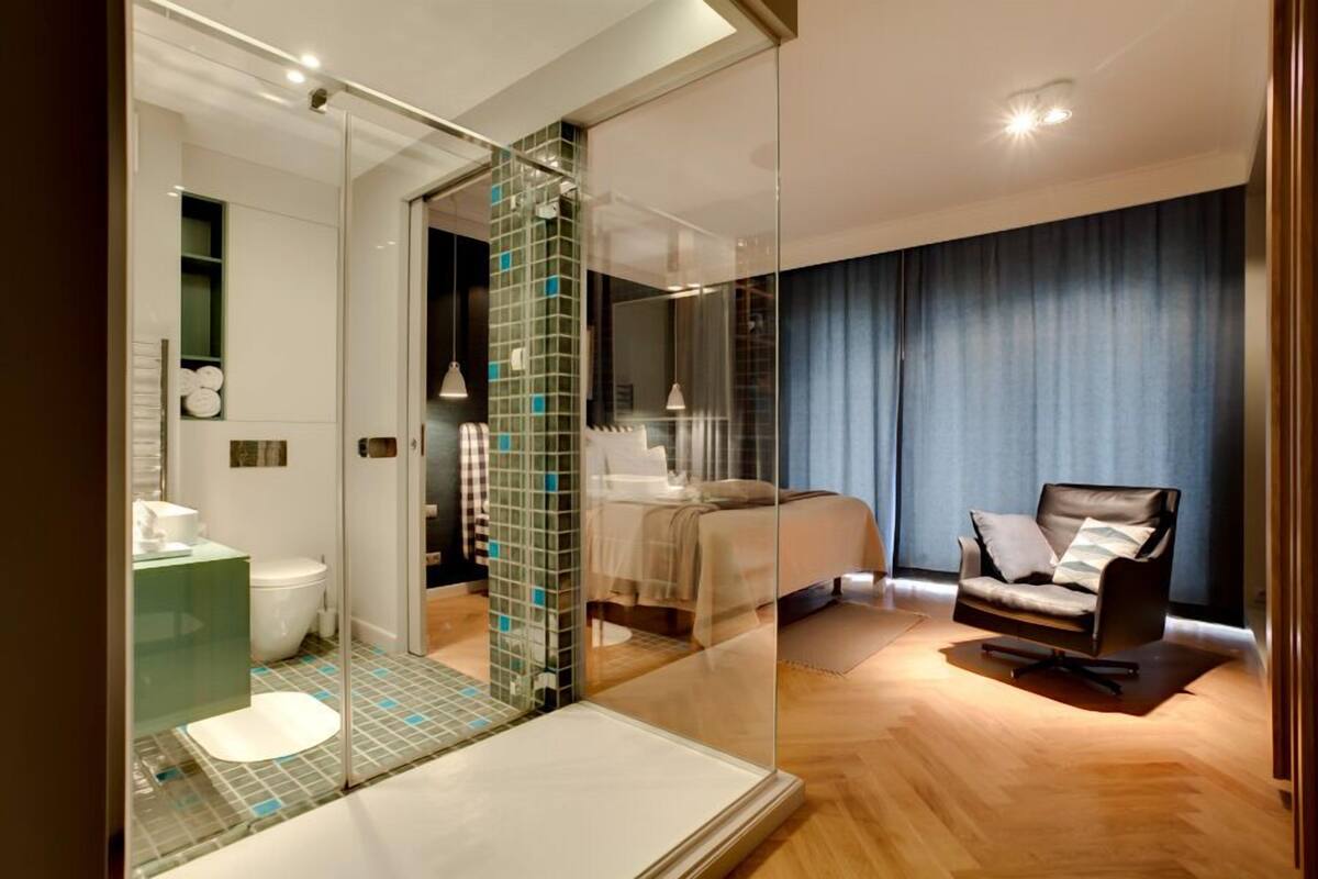 Ein modernes Schlafzimmer mit Bett, einer Sitzecke mit Sessel und einem durch eine Glasabtrennung einsehbaren Badezimmer.
