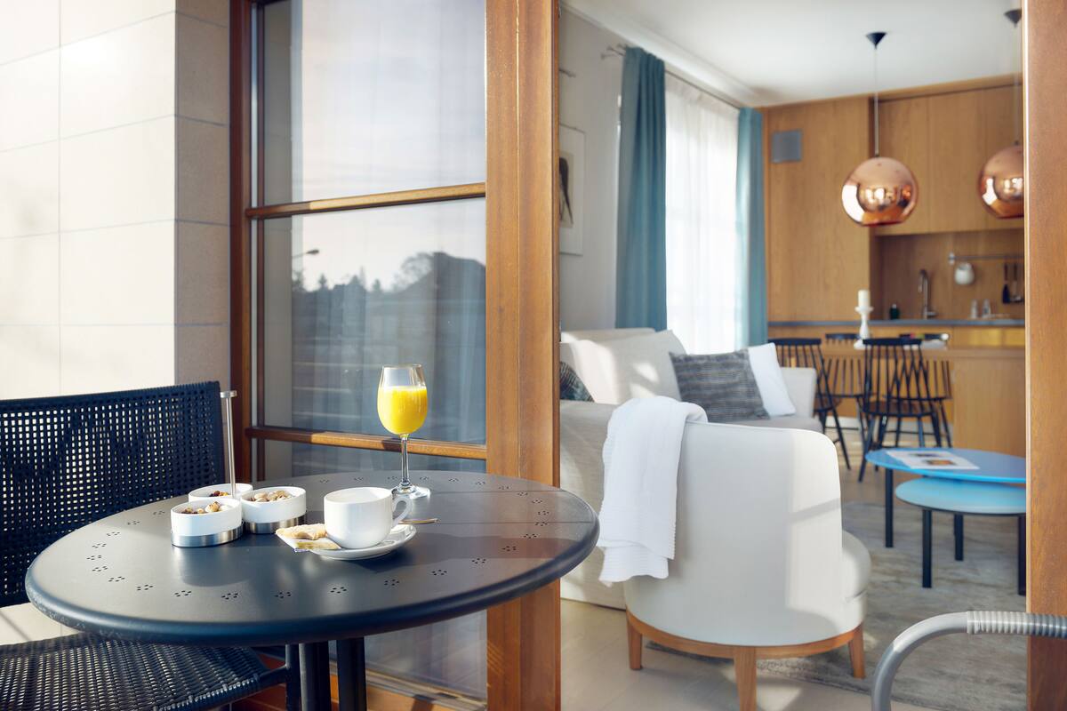 Ein modernes Hotelzimmer mit Essbereich, ausgestattet mit einem runden Tisch, Stühlen und einem Frühstücksbuffet mit Orangensaft und Schalen mit Müsli.