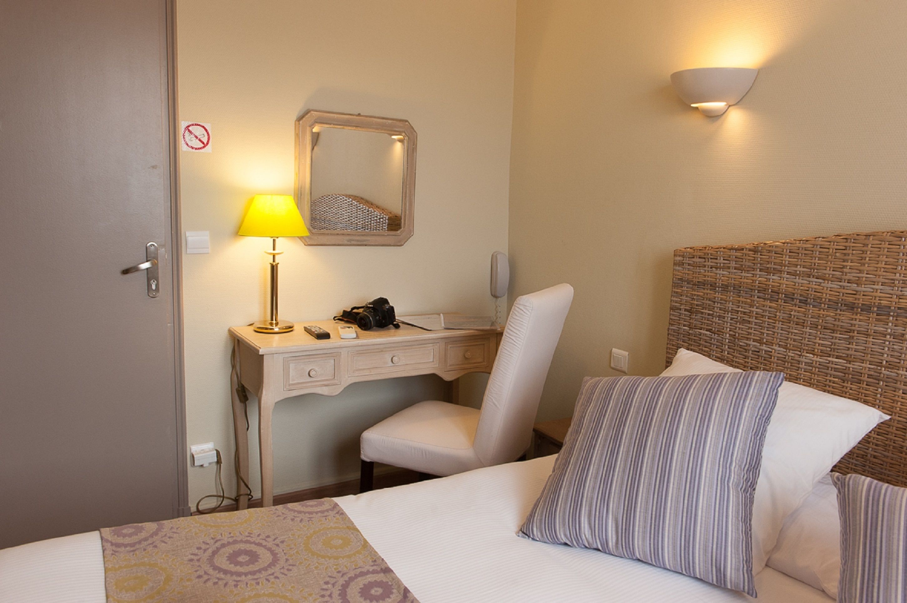 Una camera d'albergo con un letto, una scrivania con una lampada, una sedia, uno specchio e una luce a parete.