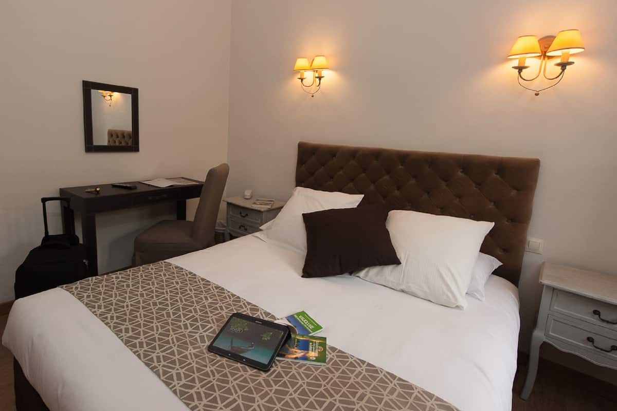 Quarto de hotel com uma cama, uma escrivaninha e duas luminárias fixadas na parede.