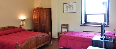 Quarto com duas camas, guarda-roupa de madeira, escrivaninha com computador e janela com luz natural.