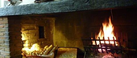 Una chimenea de ladrillo con fuego encendido, un estante de madera encima que contiene diversos utensilios de cocina y una cesta de pan sobre la repisa.