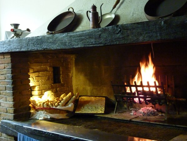 A brick fireplace with a fire burning inside, a wooden shelf above holding various kitchen utensils, and a basket of bread on the mantel.