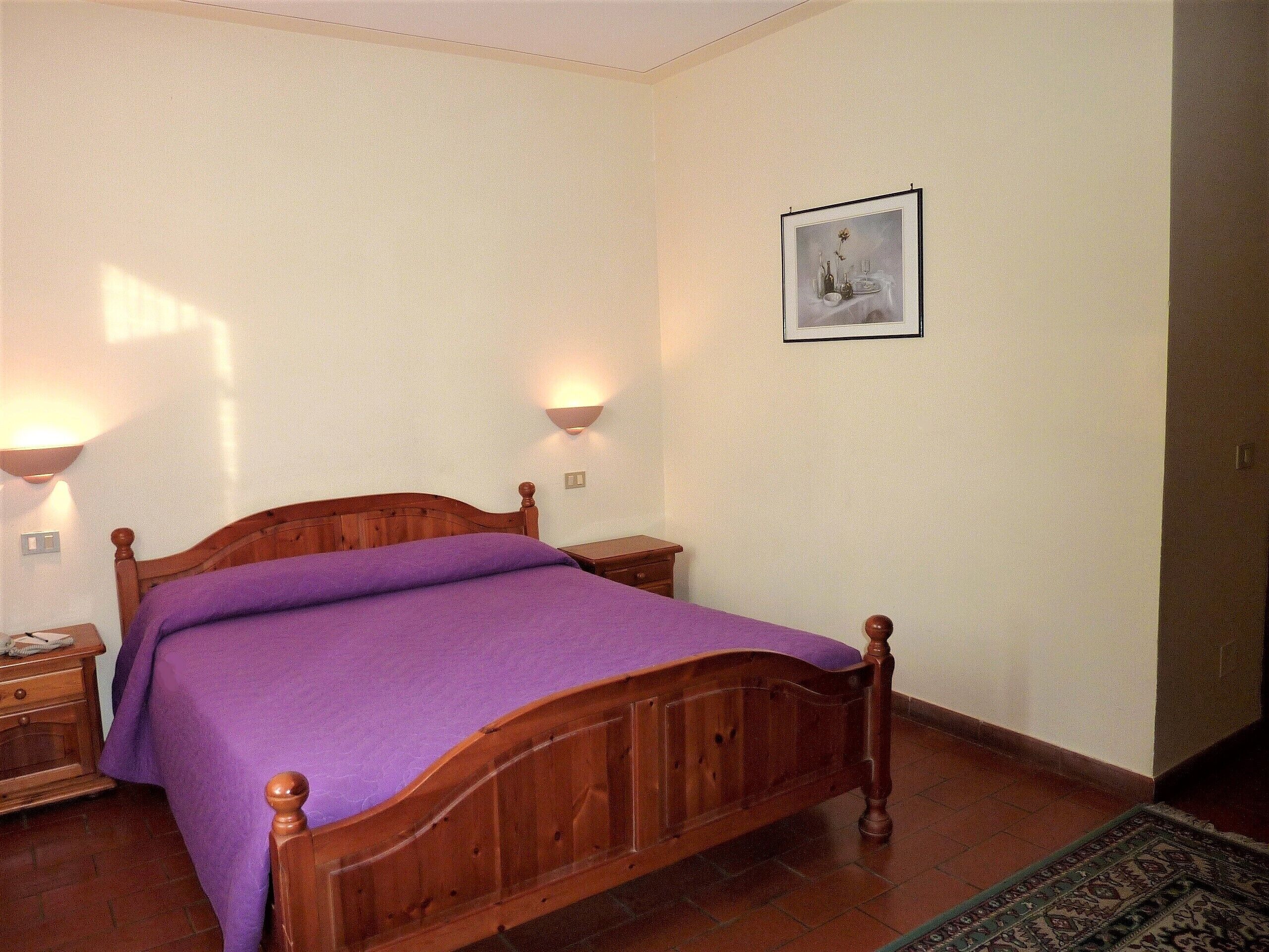 A bedroom with a wooden bed, purple bedding, two bedside tables, a nightstand, a wall-mounted lamp, and a framed picture on the wall.