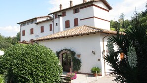 A two-story building with a tiled roof, a central entrance with a decorative arch, and a small garden area with potted plants and flowers. - Hotel La Rocca (Narni)