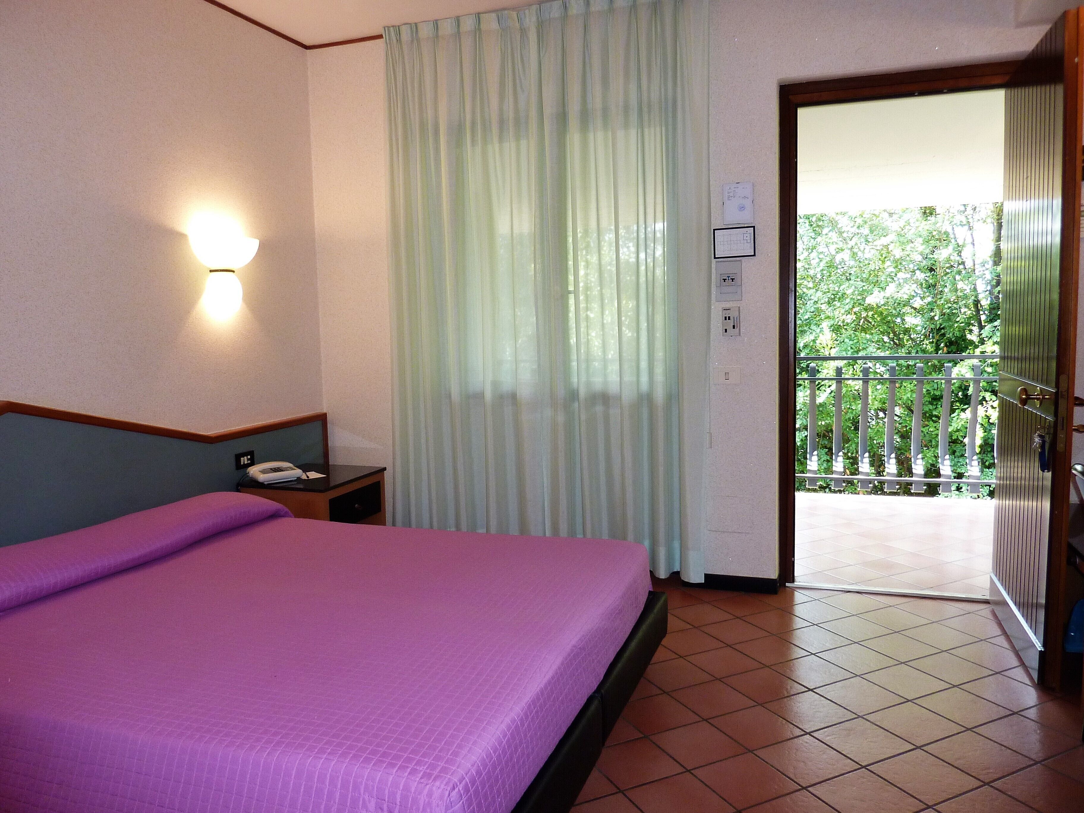 Una habitación de hotel con una cama, una mesita de noche, un teléfono, una ventana con cortinas y una puerta que da a un balcón.