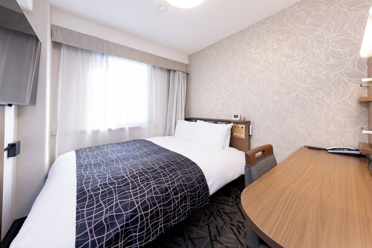 Ett hotellrum med en säng, ett skrivbord, en stol, en tv och ett fönster med gardiner.