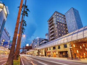 A modern hotel with a glass facade, located in a bustling urban area with tall buildings and palm trees. - APA Hotel Miyazaki Eki Tachibanadori (Miyazaki)