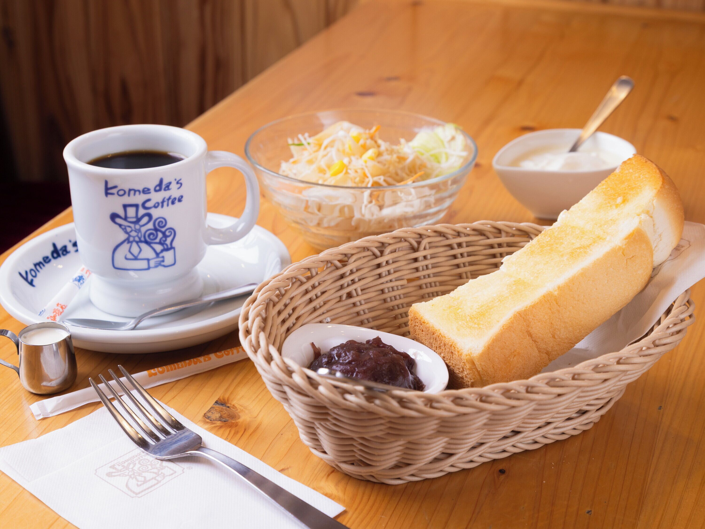 A dining table with a wicker basket containing a loaf of bread, a bowl of salad, a cup of coffee, and a spoon.