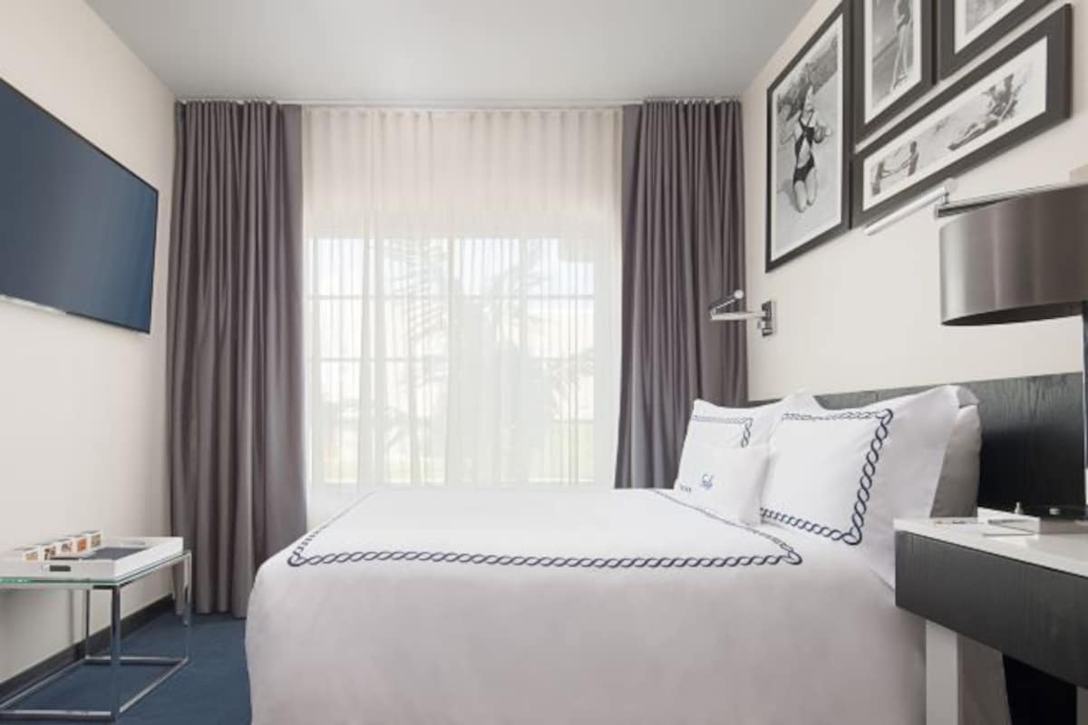 A hotel room with a bed, a desk, a chair, a TV, and a window with curtains.