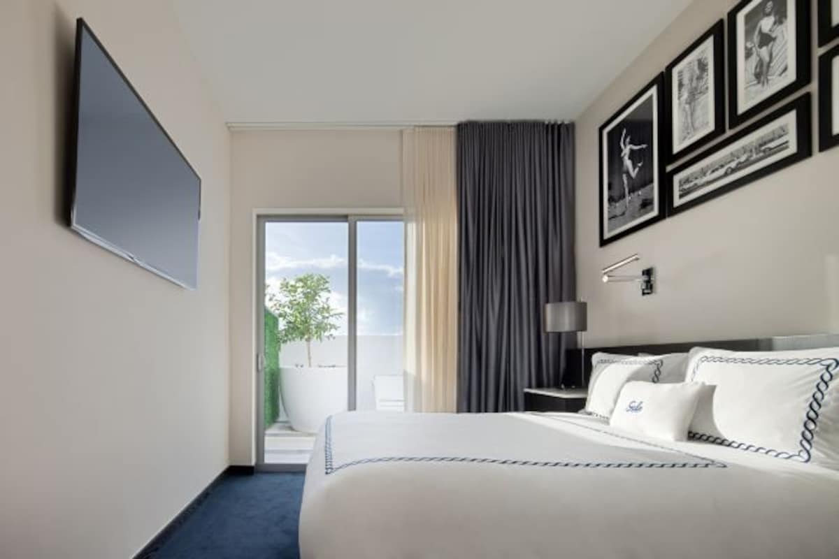 A hotel room with a bed, a flat-screen TV, a large window, and framed pictures on the wall.