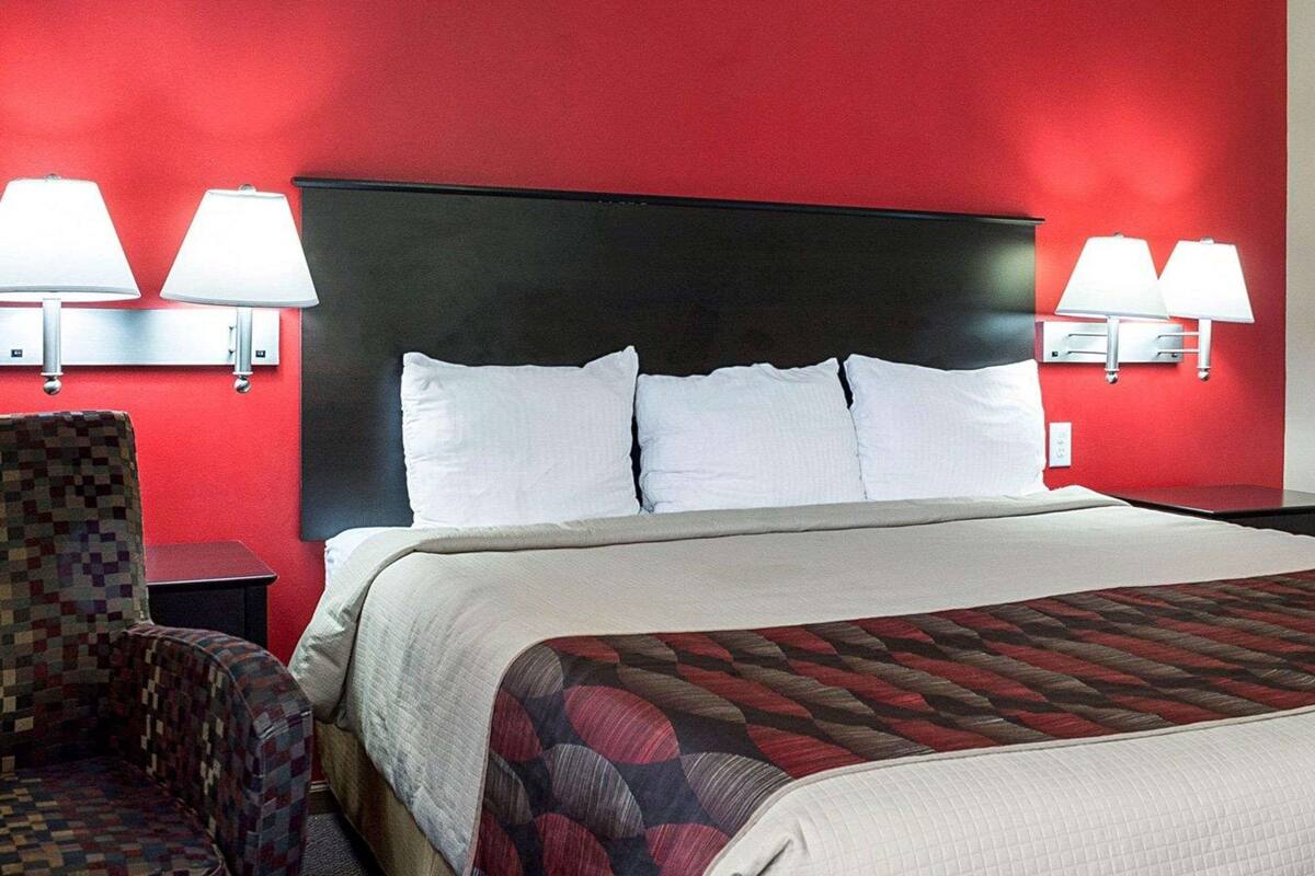 A hotel room with a red wall, a black headboard, two white pillows, a patterned throw, and two wall-mounted lamps.