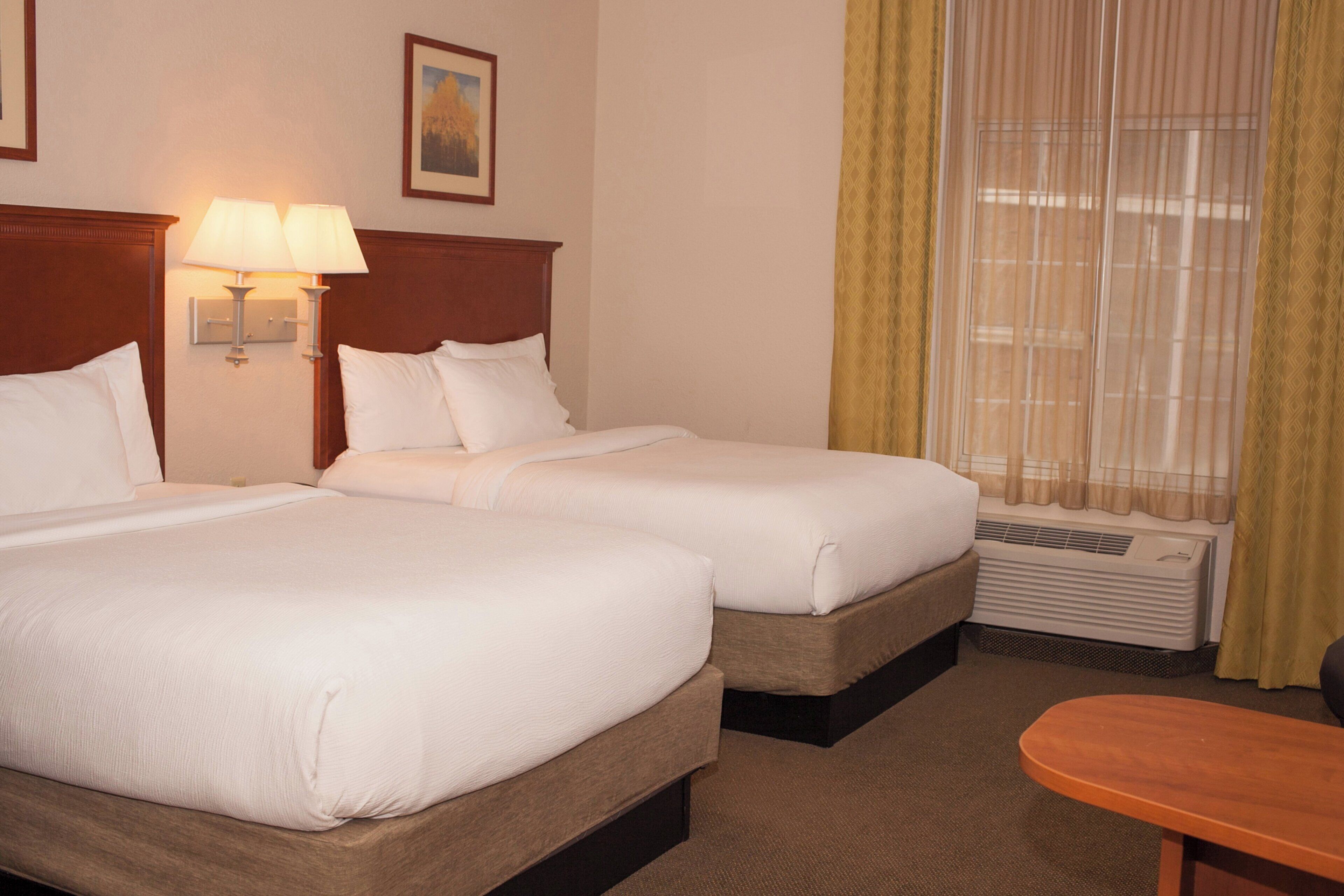 A hotel room with two beds, a nightstand with lamps, a window with curtains, and a wooden table.