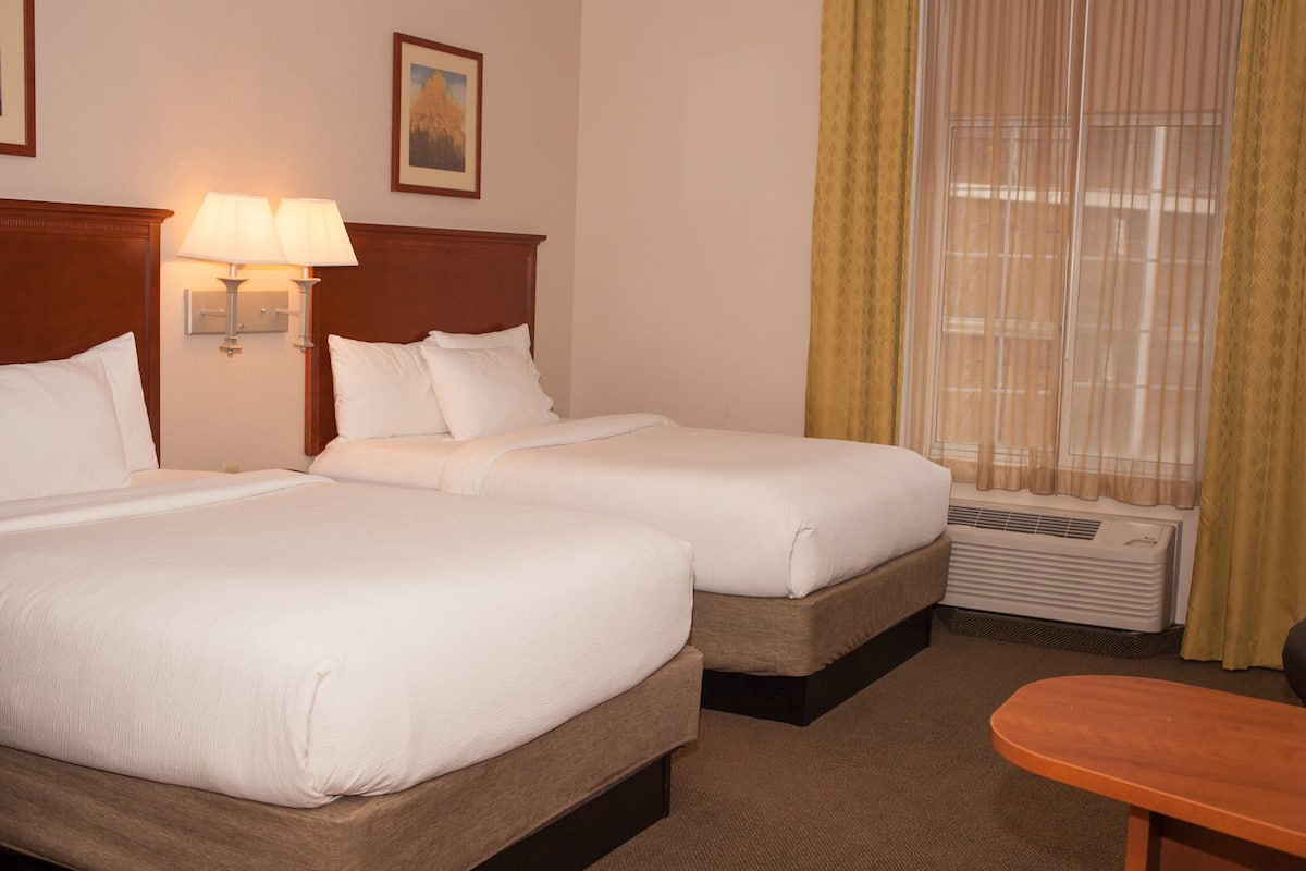 A hotel room with two beds, a nightstand with lamps, a window with curtains, and a small table.