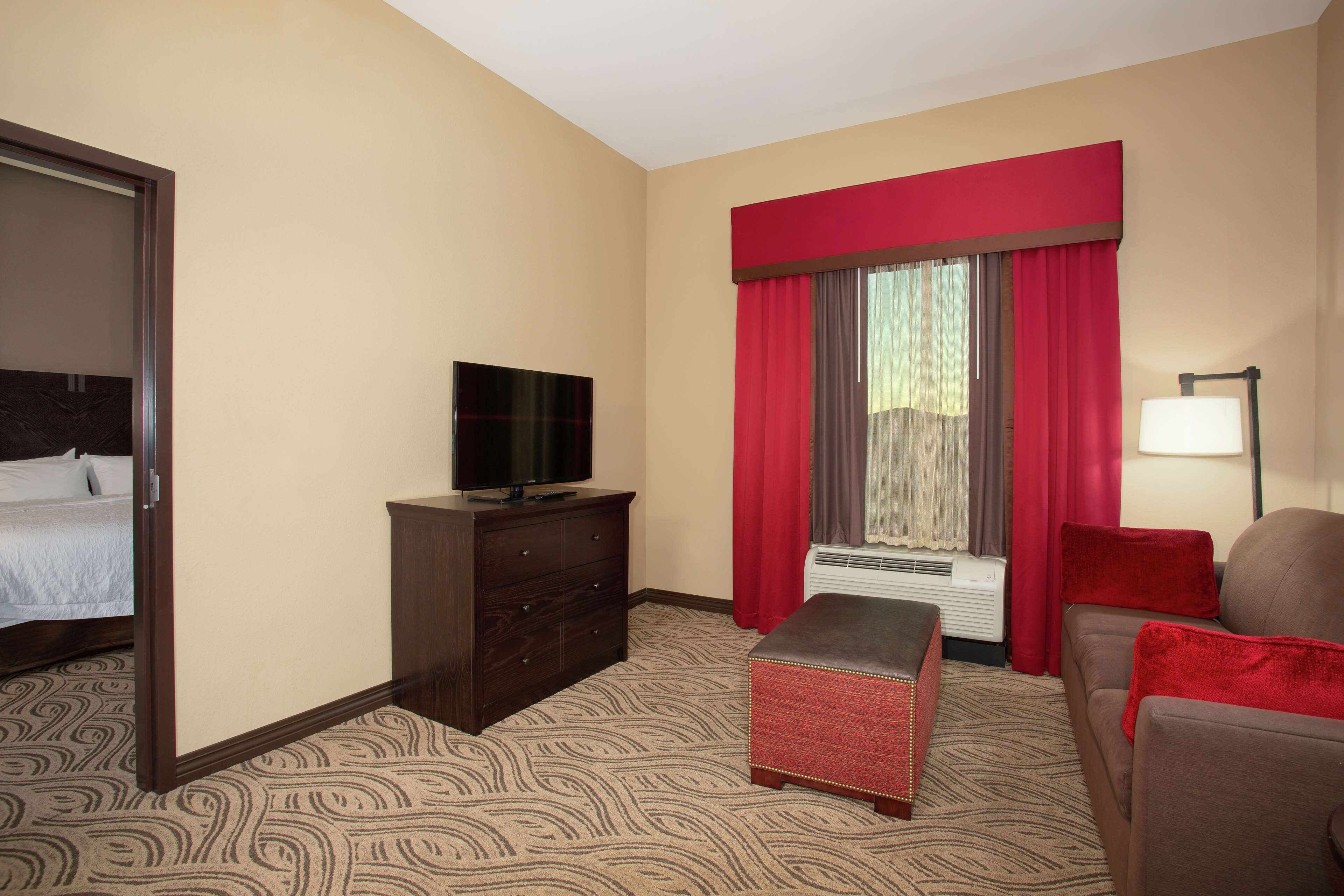 A hotel room with a bed, a TV on a dresser, a red curtain, a lamp, and a patterned carpet.