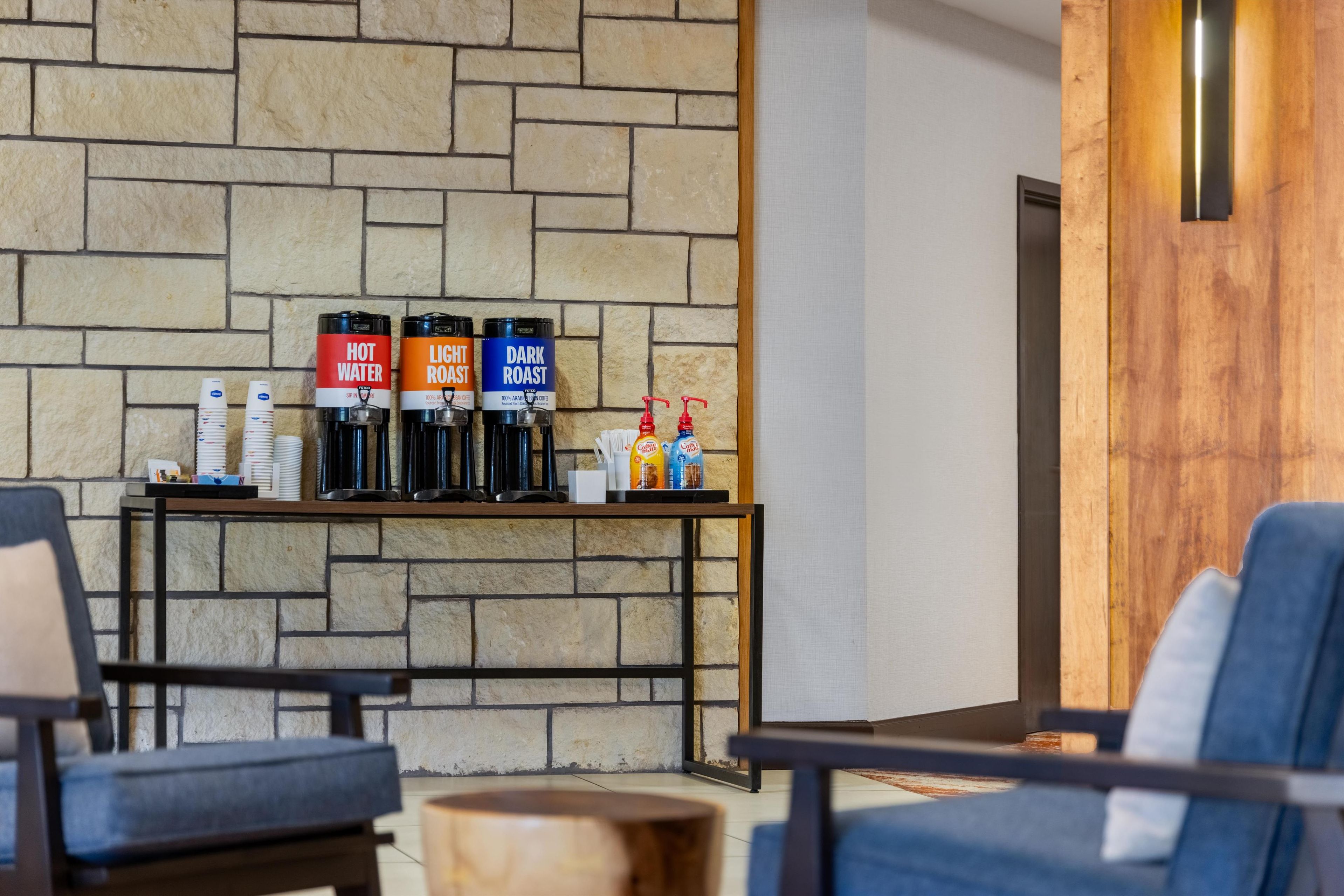 A modern lobby with a stone wall, a coffee station with labeled dispensers, and seating areas.