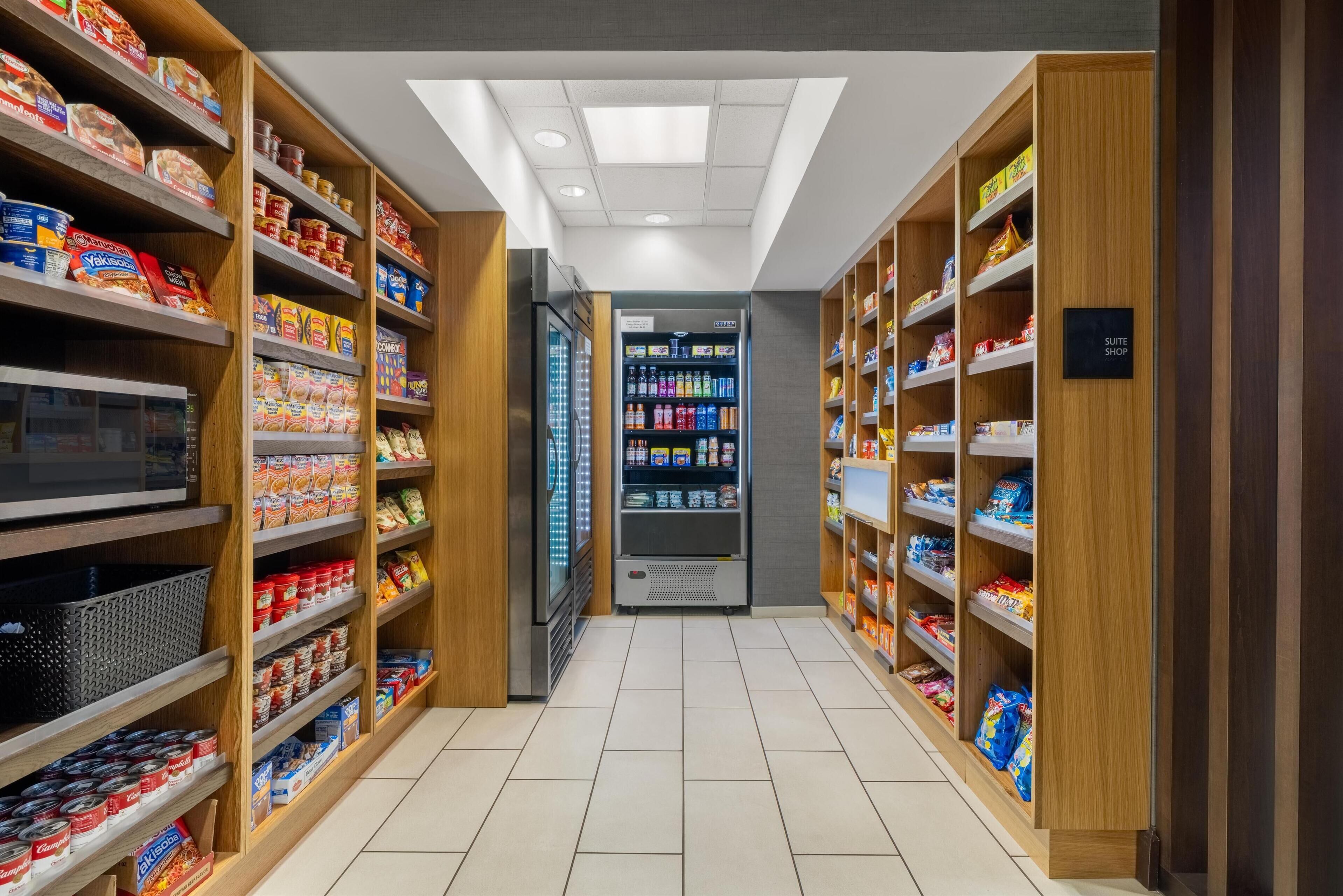 A hotel corridor with wooden shelving stocked with snacks and beverages, a refrigerator, and a vending machine.
